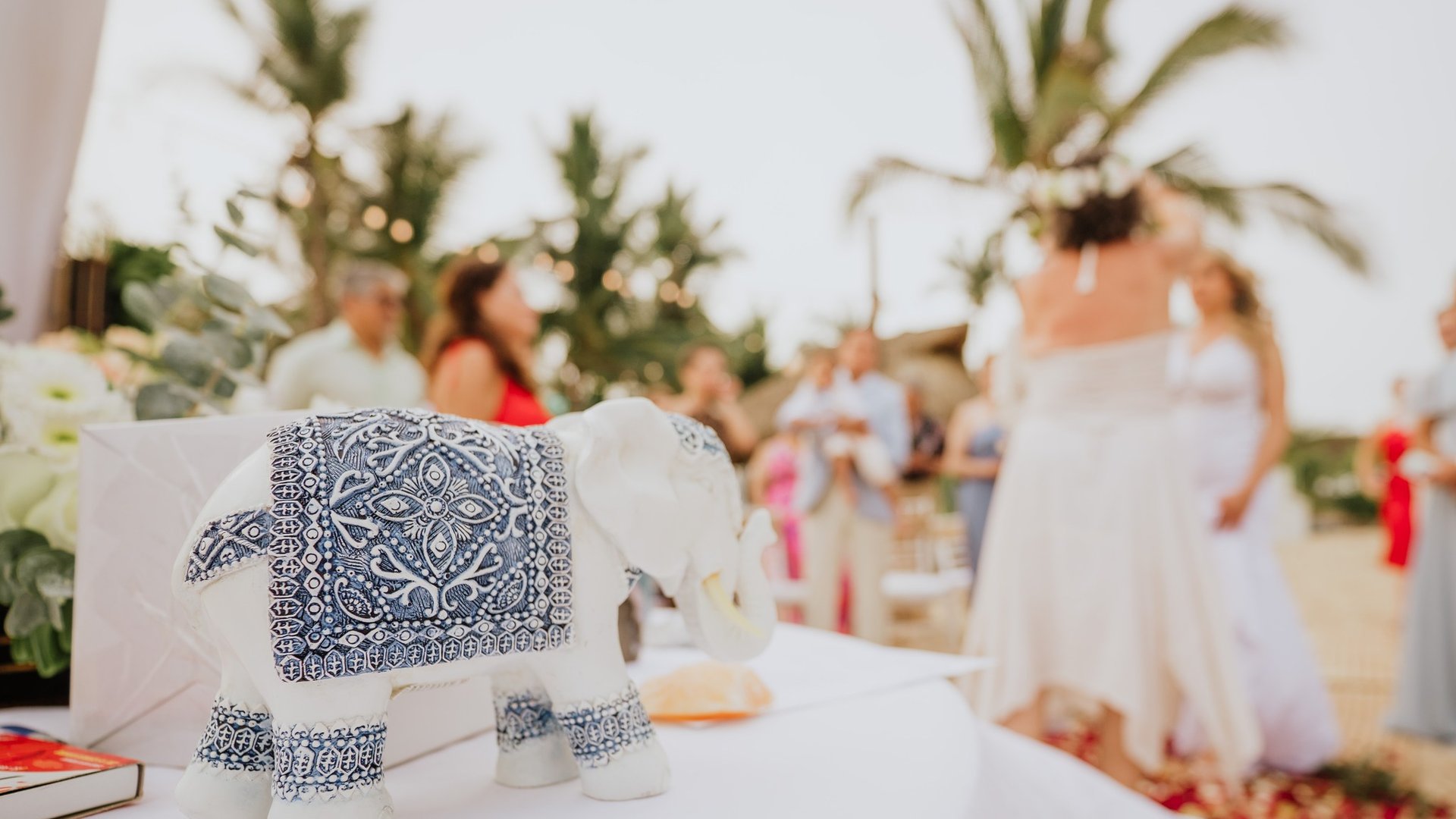 Detalle decorativo de elefante blanco en mesa de banquete con invitados y palmeras al fondo