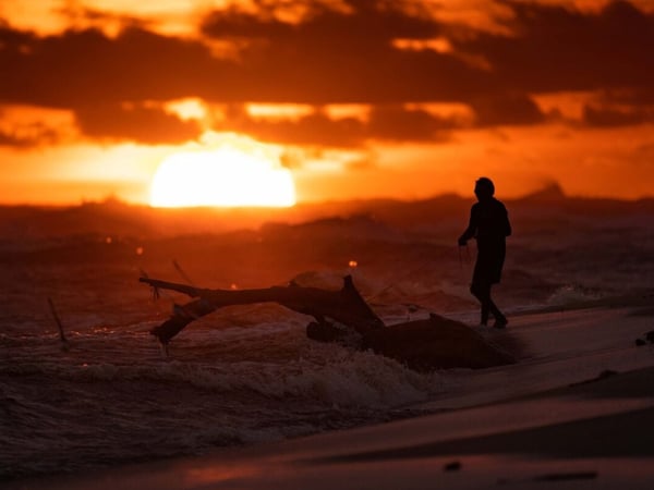 Silueta de pescador en la orilla del mar con tronco arrastrado por las olas bajo un atardecer intenso en tonos naranja y rojo