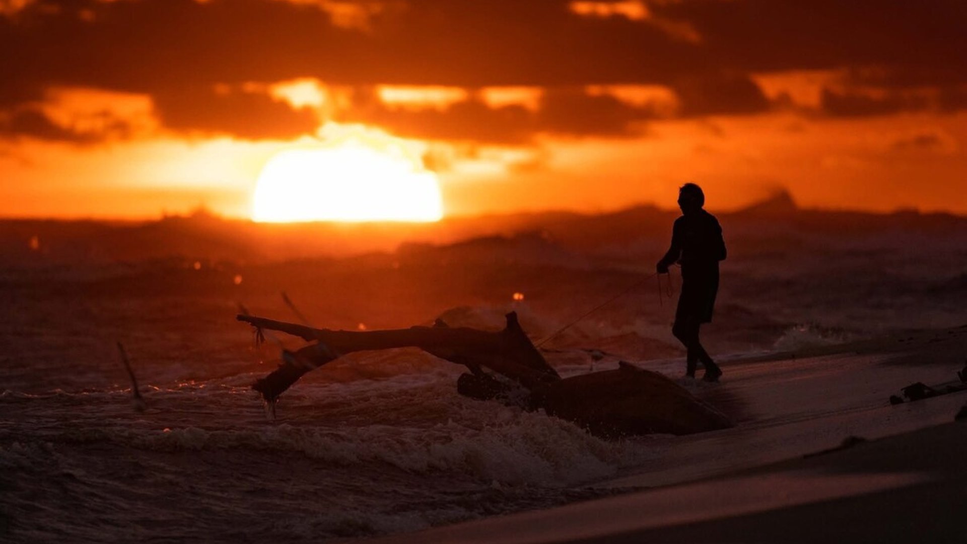 Silueta de pescador en la orilla del mar con tronco arrastrado por las olas bajo un atardecer intenso en tonos naranja y rojo