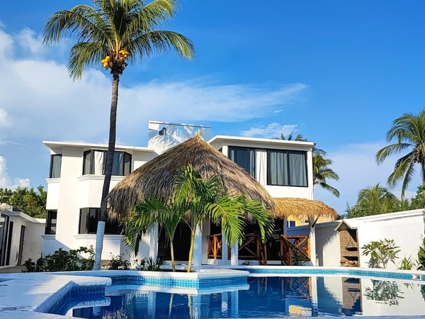 Villa blanca de dos pisos con palapa central y alberca privada bajo cielo azul despejado