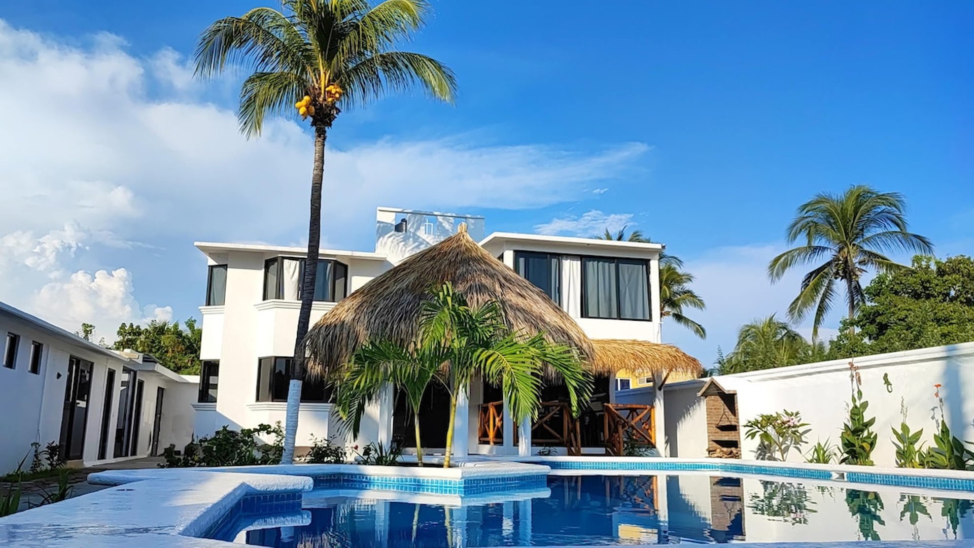Villa blanca de dos pisos con palapa central y alberca privada bajo cielo azul despejado
