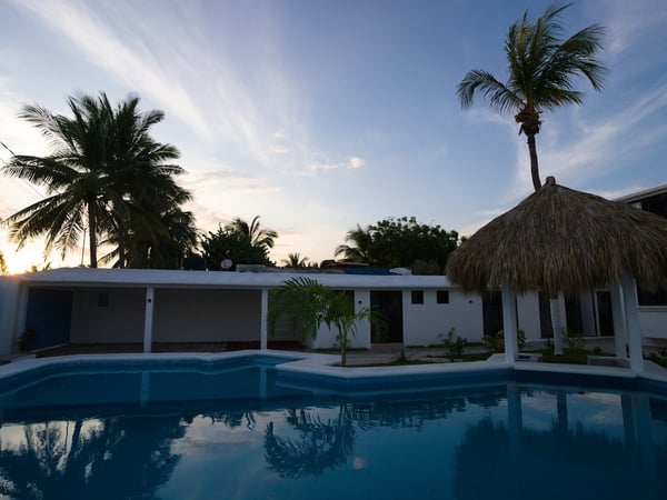 Alberca amplia al atardecer con palapa y construcción blanca al fondo, palmeras enmarcando el cielo crepuscular
