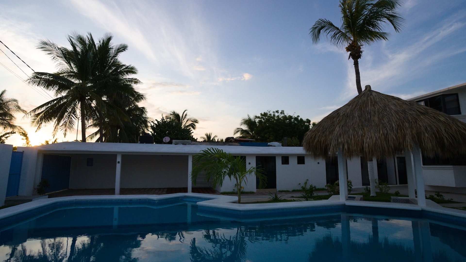 Alberca amplia al atardecer con palapa y construcción blanca al fondo, palmeras enmarcando el cielo crepuscular