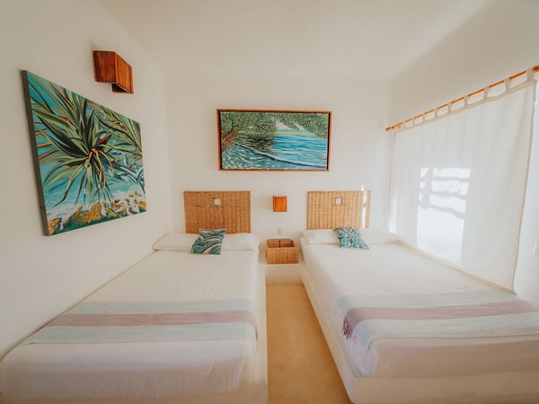 Recámara con dos camas individuales, cabeceras de mimbre y pinturas tropicales en paredes blancas