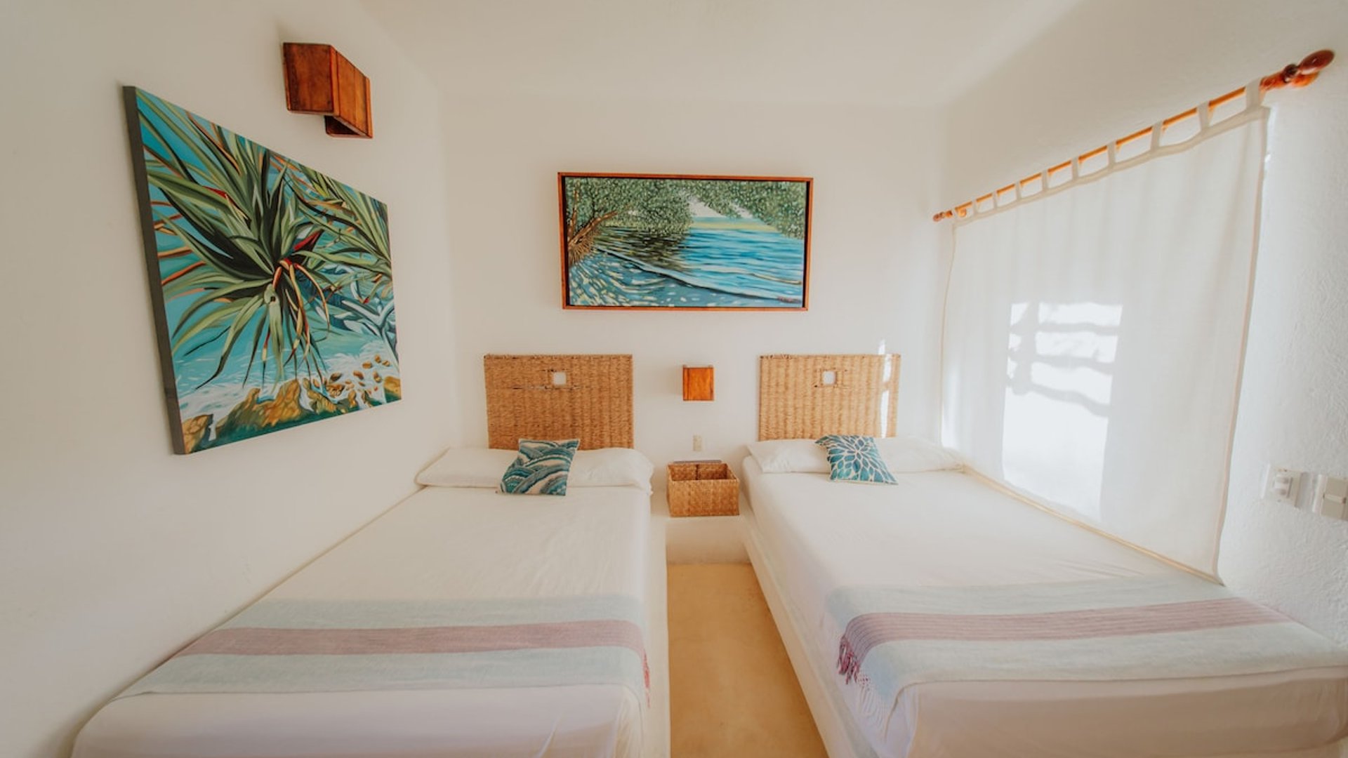 Recámara con dos camas individuales, cabeceras de mimbre y pinturas tropicales en paredes blancas