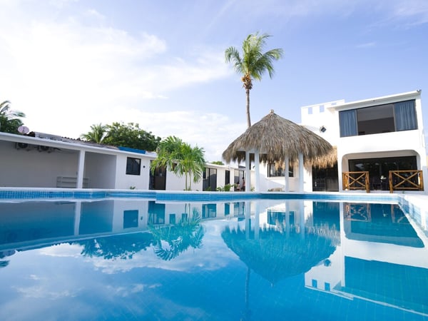 Alberca de mosaico azul frente a palapa circular y villa blanca de dos niveles bajo cielo despejado