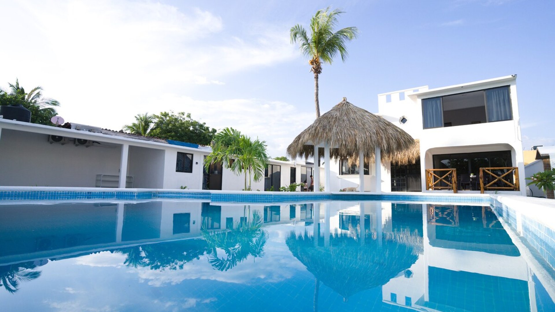 Alberca de mosaico azul frente a palapa circular y villa blanca de dos niveles bajo cielo despejado
