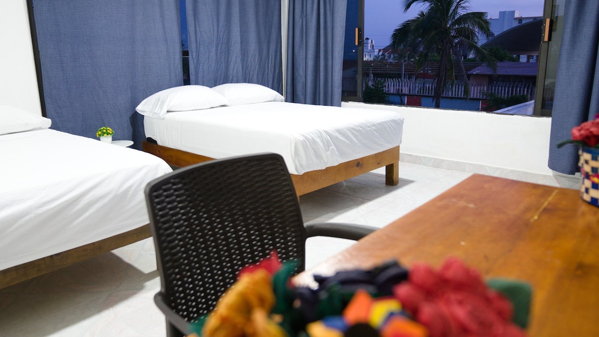 Recámara con dos camas individuales, cortinas azules y ventana amplia con vista nocturna a palmeras y tejados