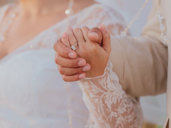 Manos entrelazadas de recién casados mostrando anillo de compromiso con velo de novia al fondo