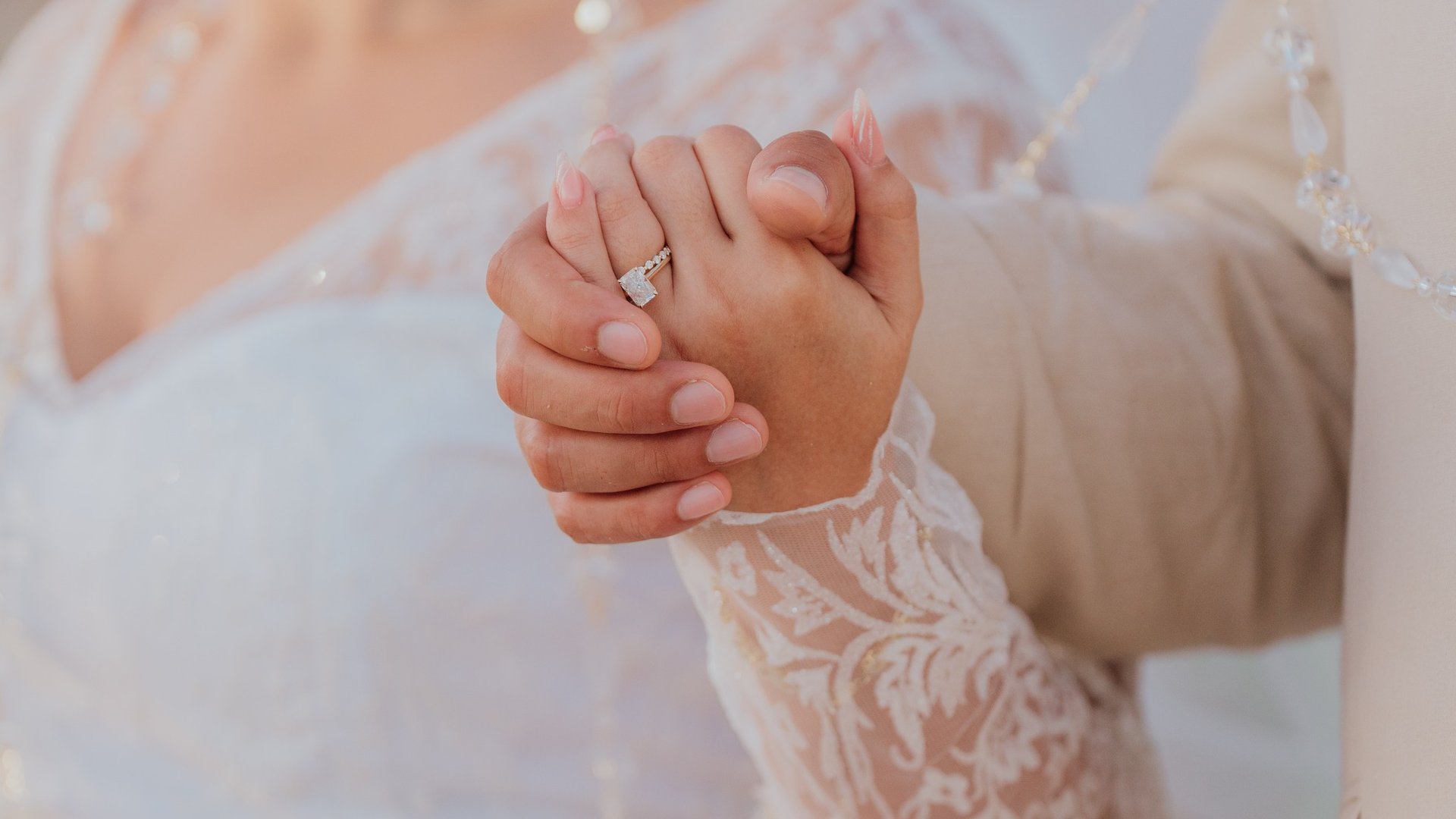 Manos entrelazadas de recién casados mostrando anillo de compromiso con velo de novia al fondo
