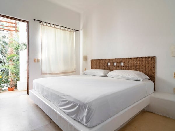 Recámara blanca con cama doble, cabecera de mimbre y puerta abierta hacia jardín tropical