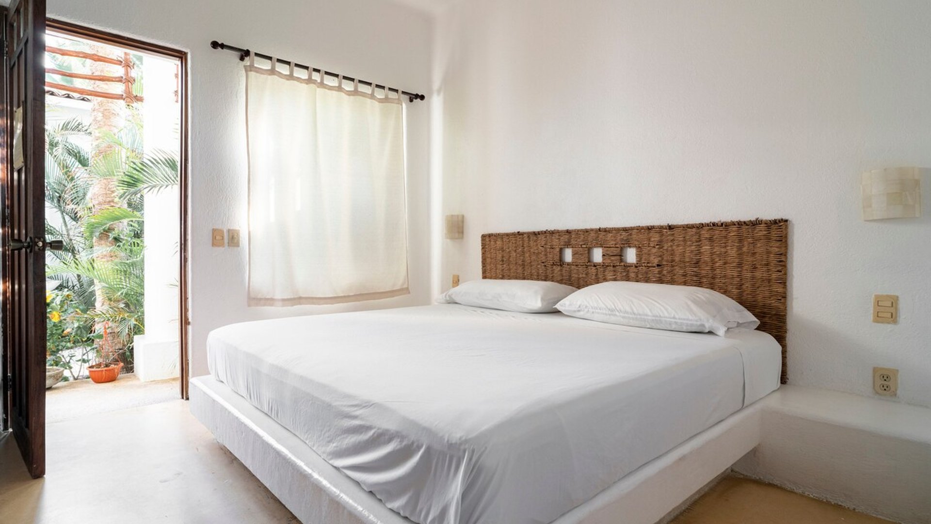 Recámara blanca con cama doble, cabecera de mimbre y puerta abierta hacia jardín tropical