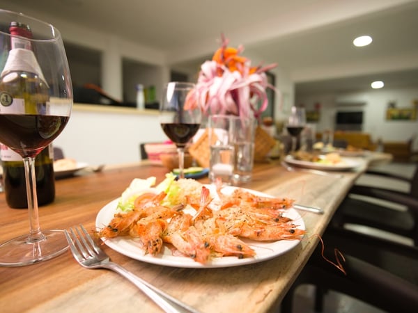 Mesa del comedor con plato de camarones al ajillo, copas de vino tinto y arreglo floral, sala al fondo