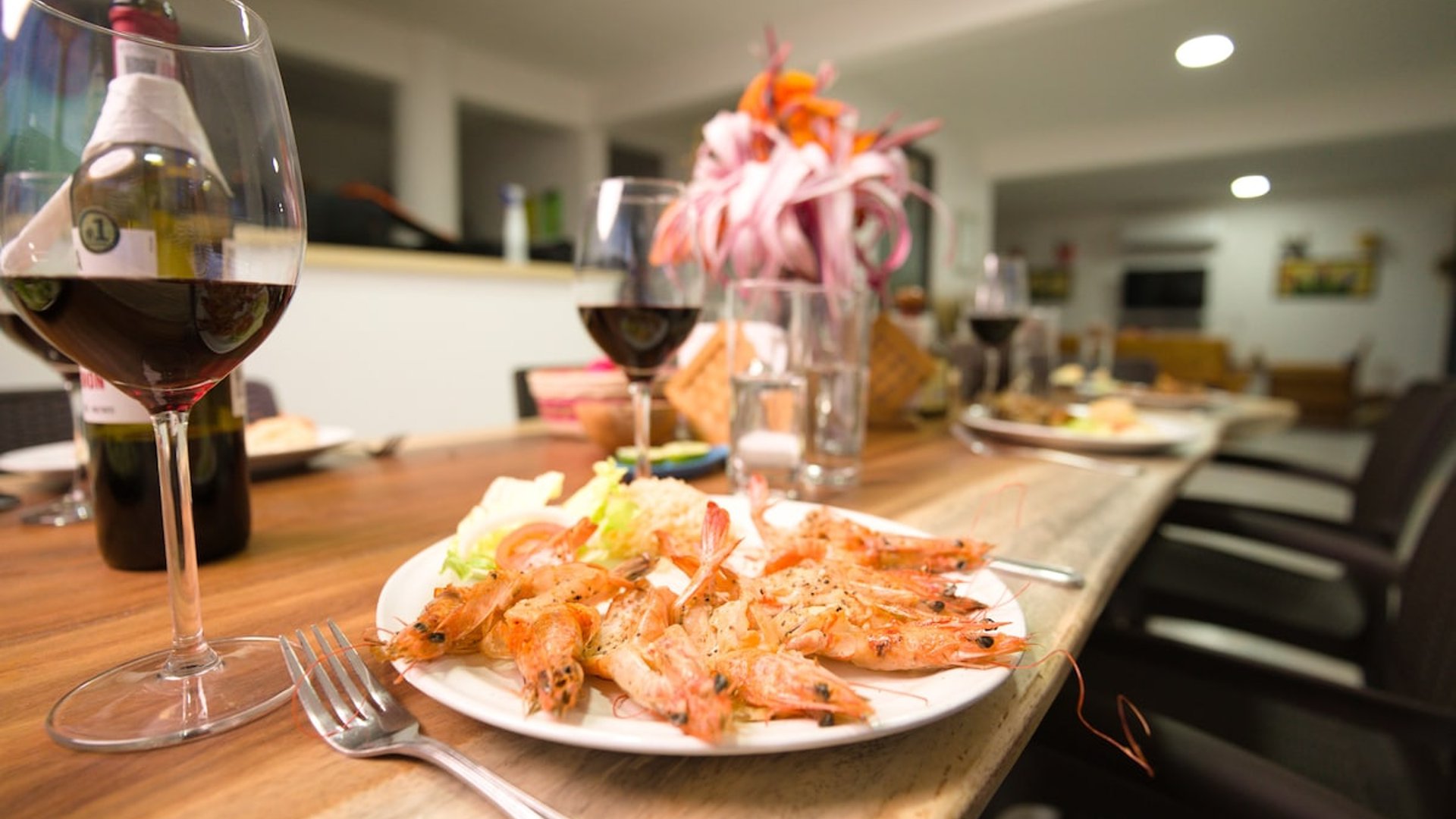 Mesa del comedor con plato de camarones al ajillo, copas de vino tinto y arreglo floral, sala al fondo