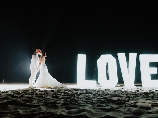 Pareja de novios besándose en la playa de noche junto a letras luminosas gigantes que forman la palabra LOVE