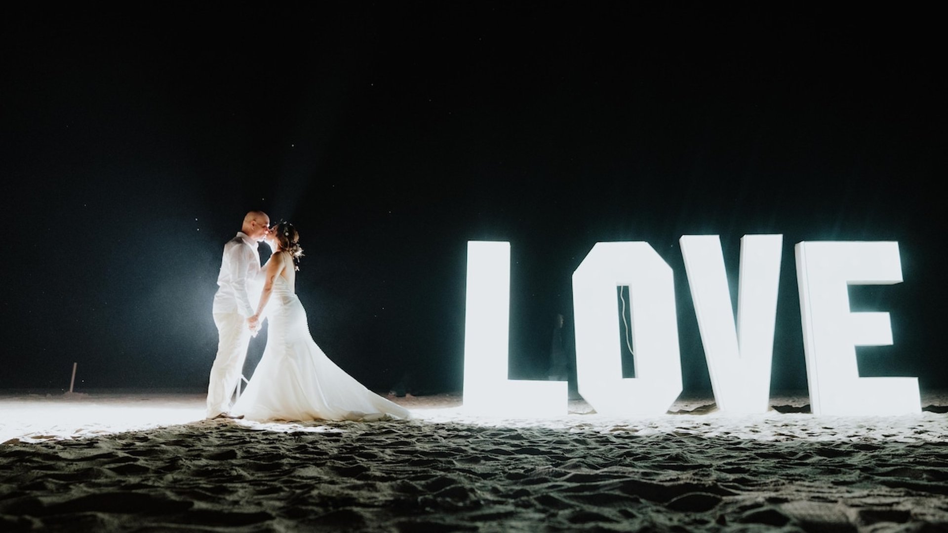 Pareja de novios besándose en la playa de noche junto a letras luminosas gigantes que forman la palabra LOVE