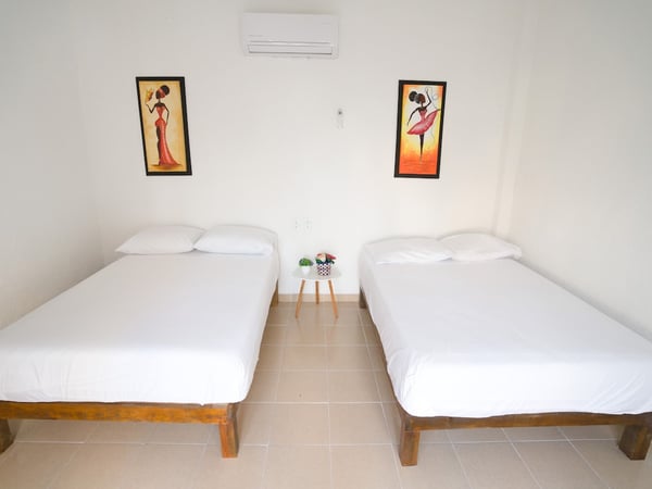 Recámara con dos camas matrimoniales de madera, ropa blanca impecable, aire acondicionado y cuadros coloridos en pared blanca