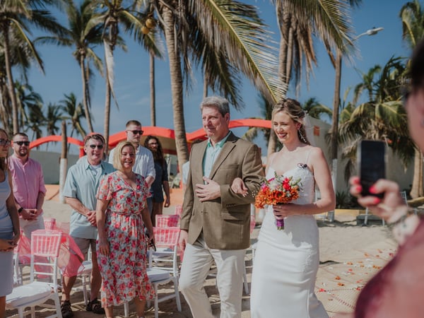 Entrada de novia en ceremonia playera con palmeras y sombrillas coral al fondo