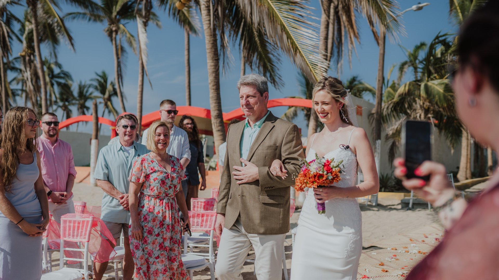 Entrada de novia en ceremonia playera con palmeras y sombrillas coral al fondo