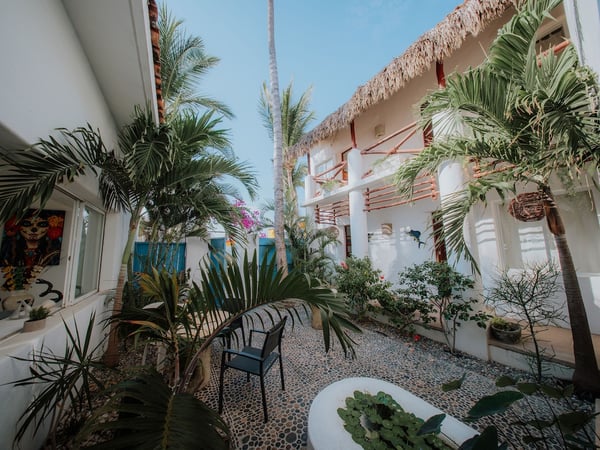 Patio interior con jardín tropical, palmeras y edificio de dos pisos con techo de palapa bajo cielo azul