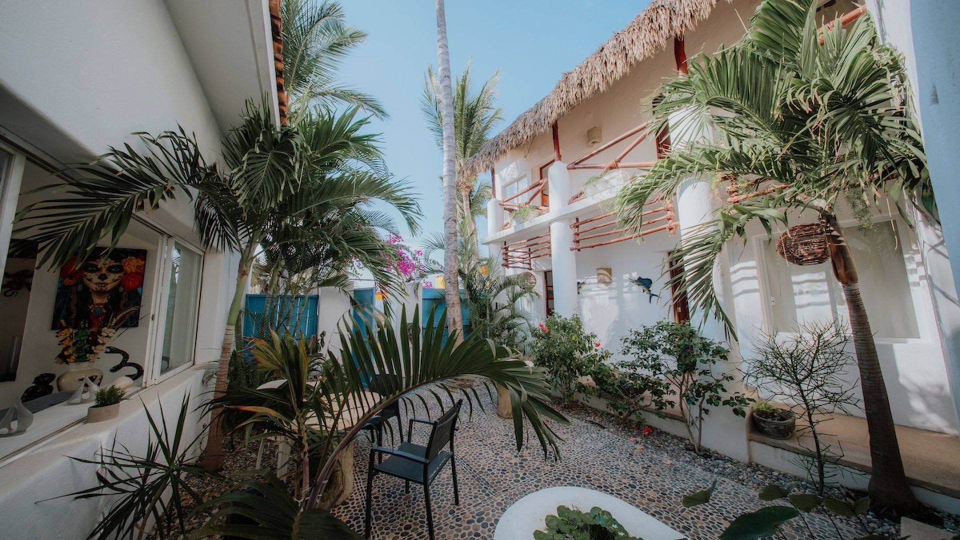 Patio interior con jardín tropical, palmeras y edificio de dos pisos con techo de palapa bajo cielo azul