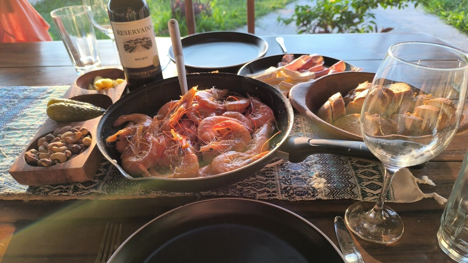 Mesa exterior con camarones al ajillo en sartén, vino tinto, pan y botanas bajo luz cálida de tarde