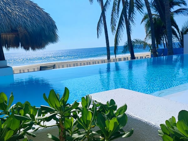 Alberca de borde infinito con vista al mar, palmeras y palapa bajo cielo azul brillante