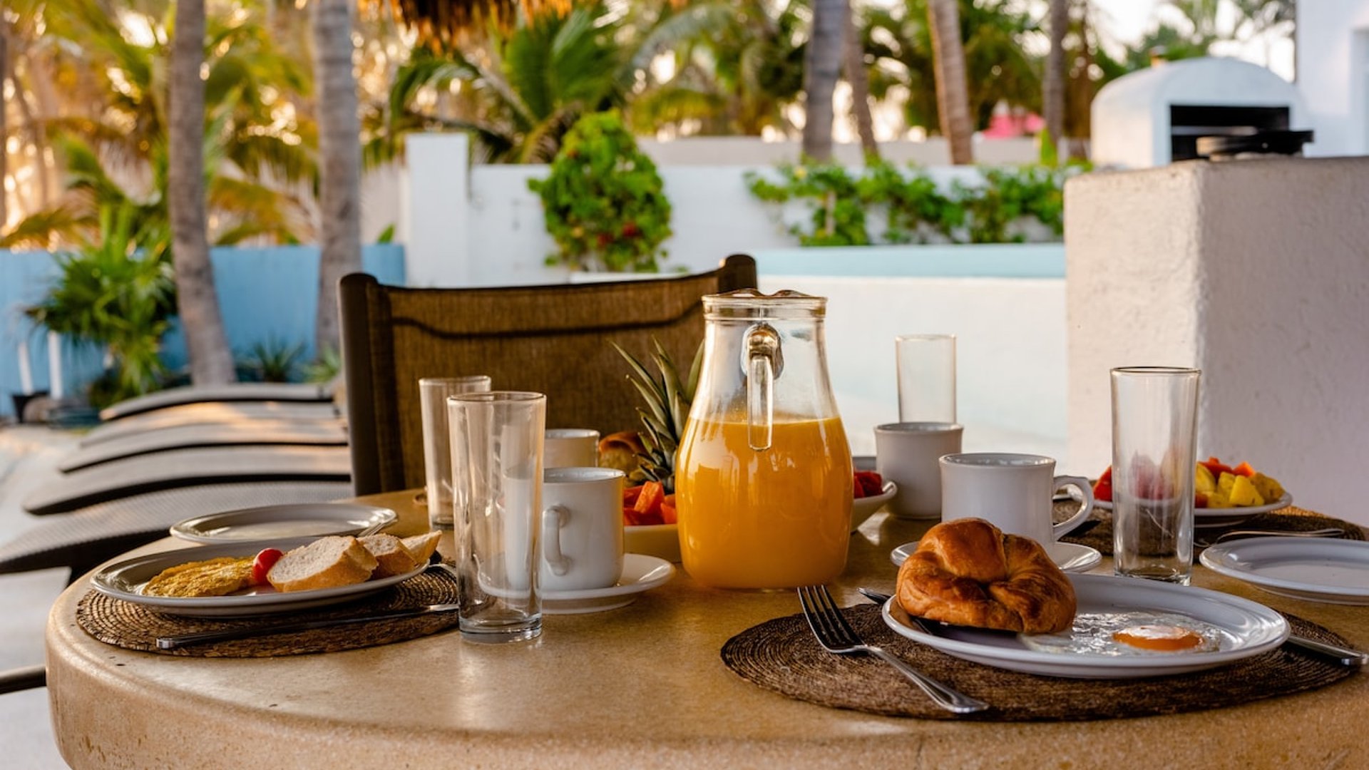 Mesa de desayuno exterior bajo palapa con jugo de naranja, croissants y frutas frescas, rodeada de palmeras