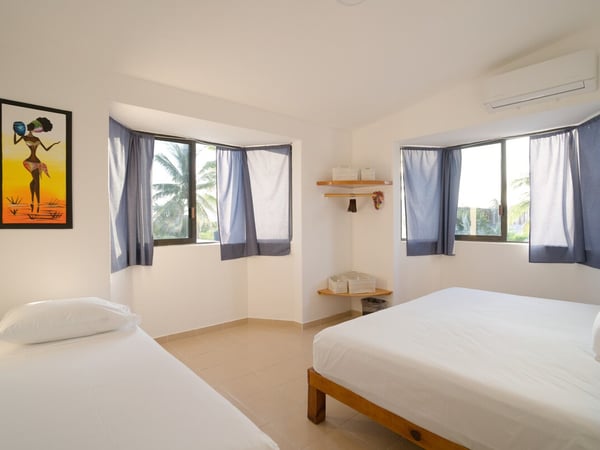 Recámara amplia y luminosa con dos camas, paredes blancas, cortinas azules y vista a palmeras desde las ventanas