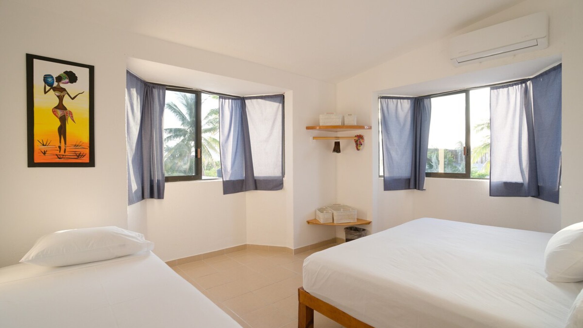 Recámara amplia y luminosa con dos camas, paredes blancas, cortinas azules y vista a palmeras desde las ventanas