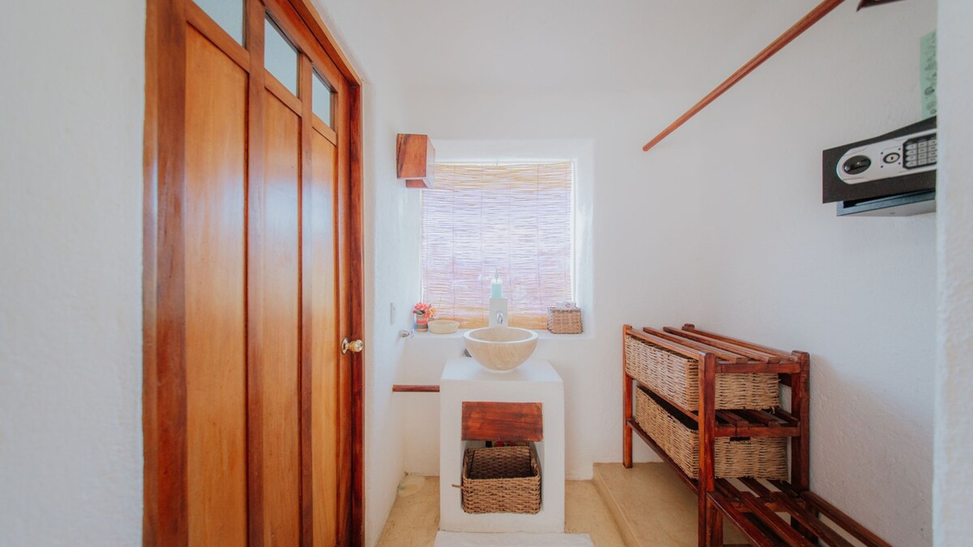 Área de lavabo con tazón de piedra sobre base blanca, mueble de mimbre y puerta de madera con ventana de bambú