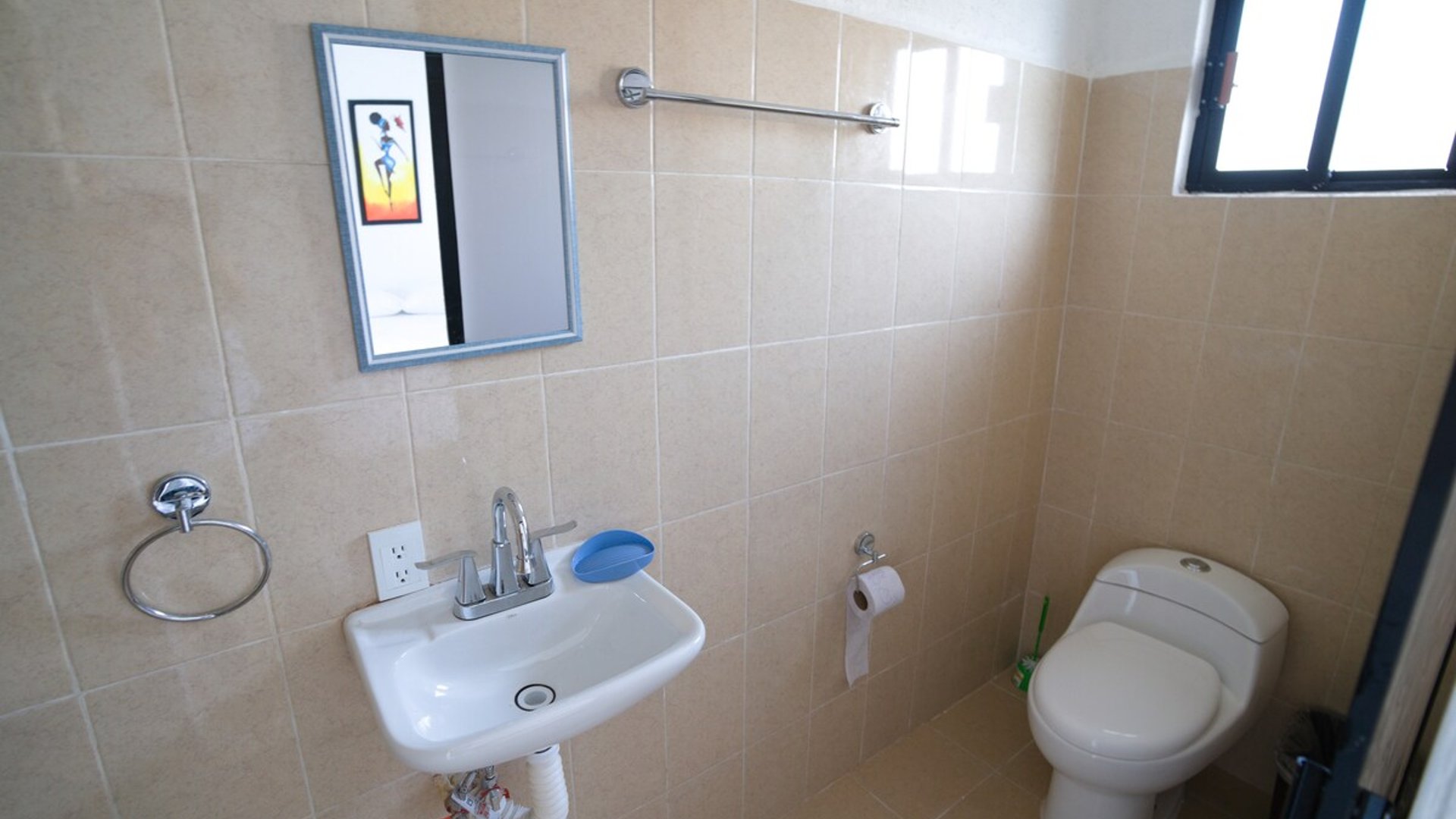 Baño sencillo con lavabo, espejo y sanitario, revestido de azulejo beige y accesorios cromados