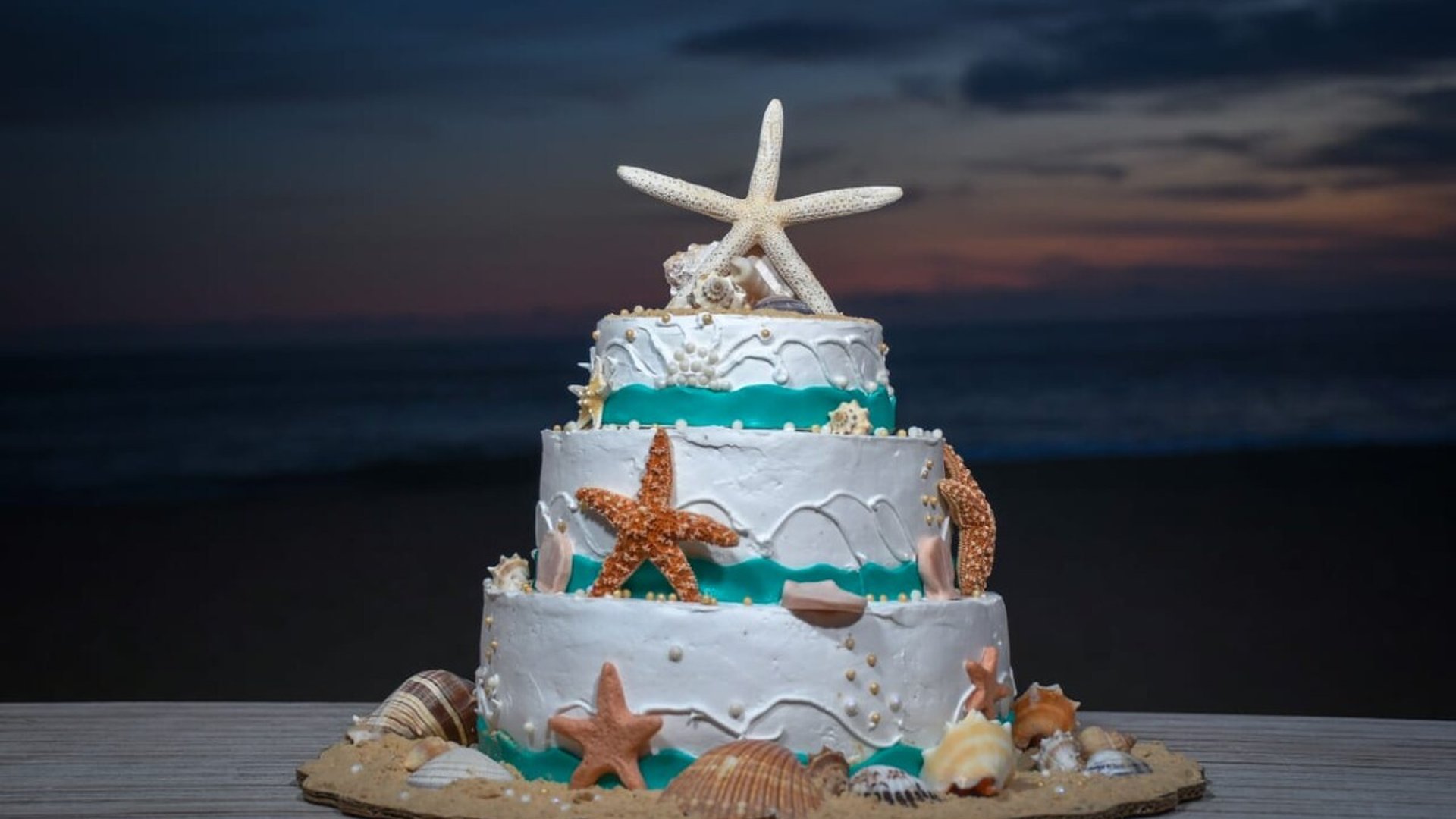 Pastel de bodas de tres pisos decorado con estrellas de mar y conchas frente al mar al atardecer