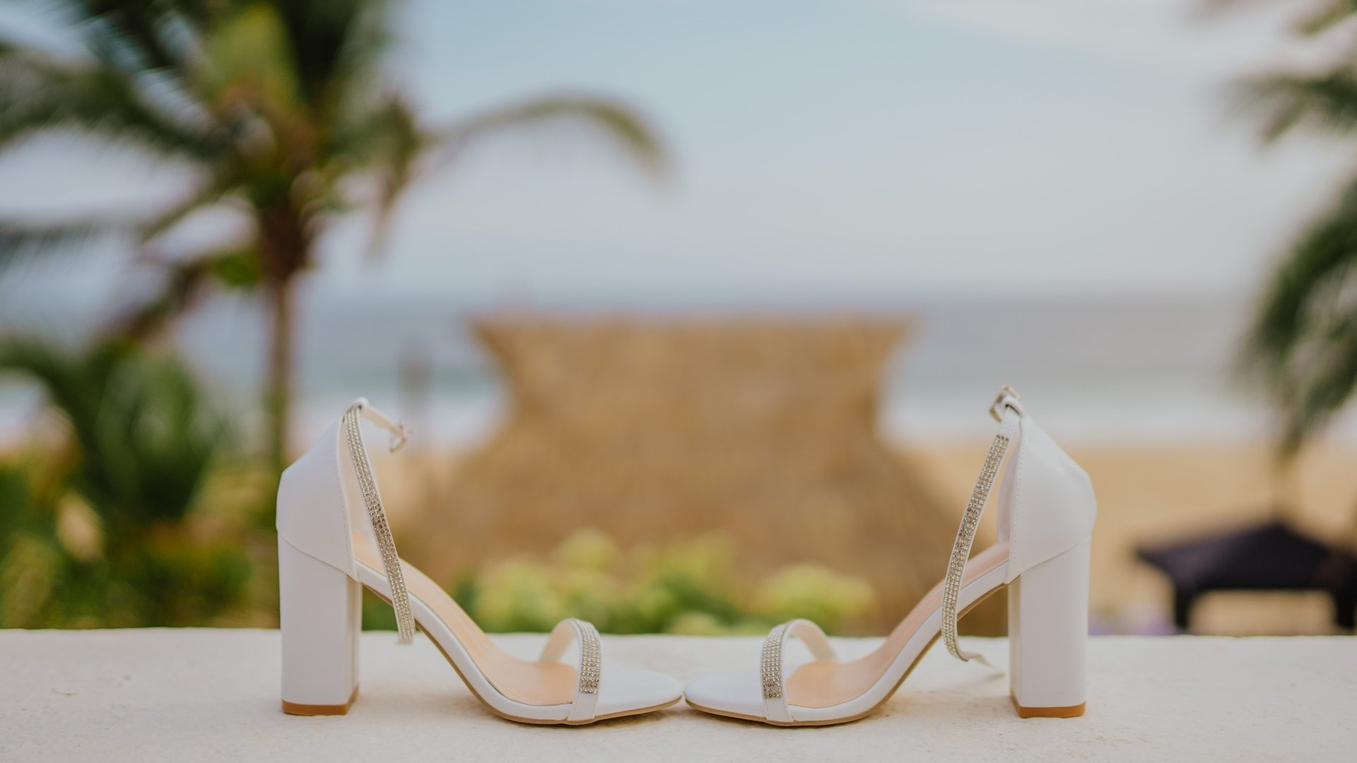 Zapatos blancos de tacón con detalles brillantes sobre muro con vista al Pacífico y palmeras