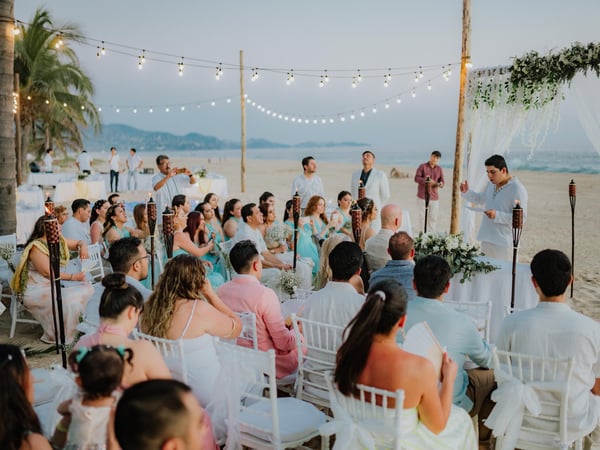 Ceremonia de boda en playa con arco floral blanco, sillas para invitados y luces colgantes al atardecer