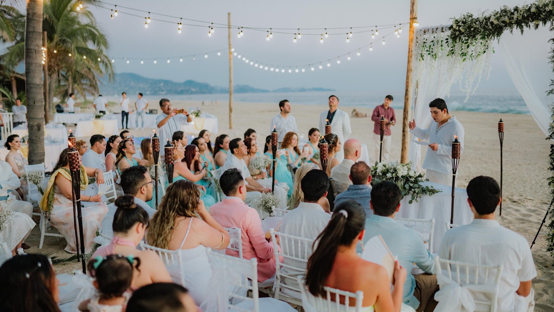 Ceremonia de boda en playa con arco floral blanco, sillas para invitados y luces colgantes al atardecer