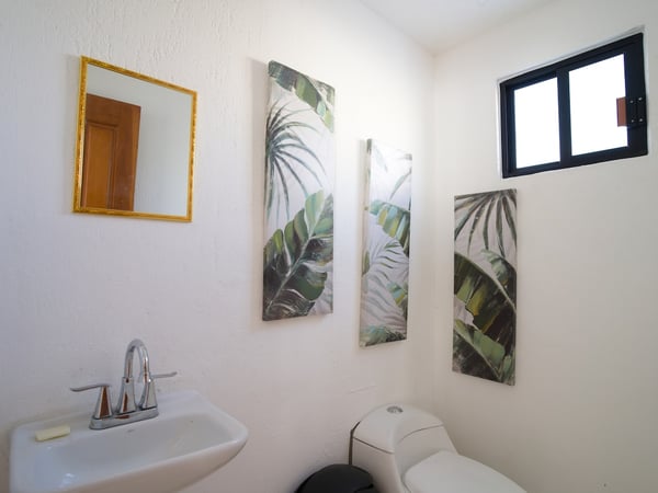 Baño blanco con lavabo, espejo dorado y tres cuadros decorativos de hojas tropicales en tonos verdes