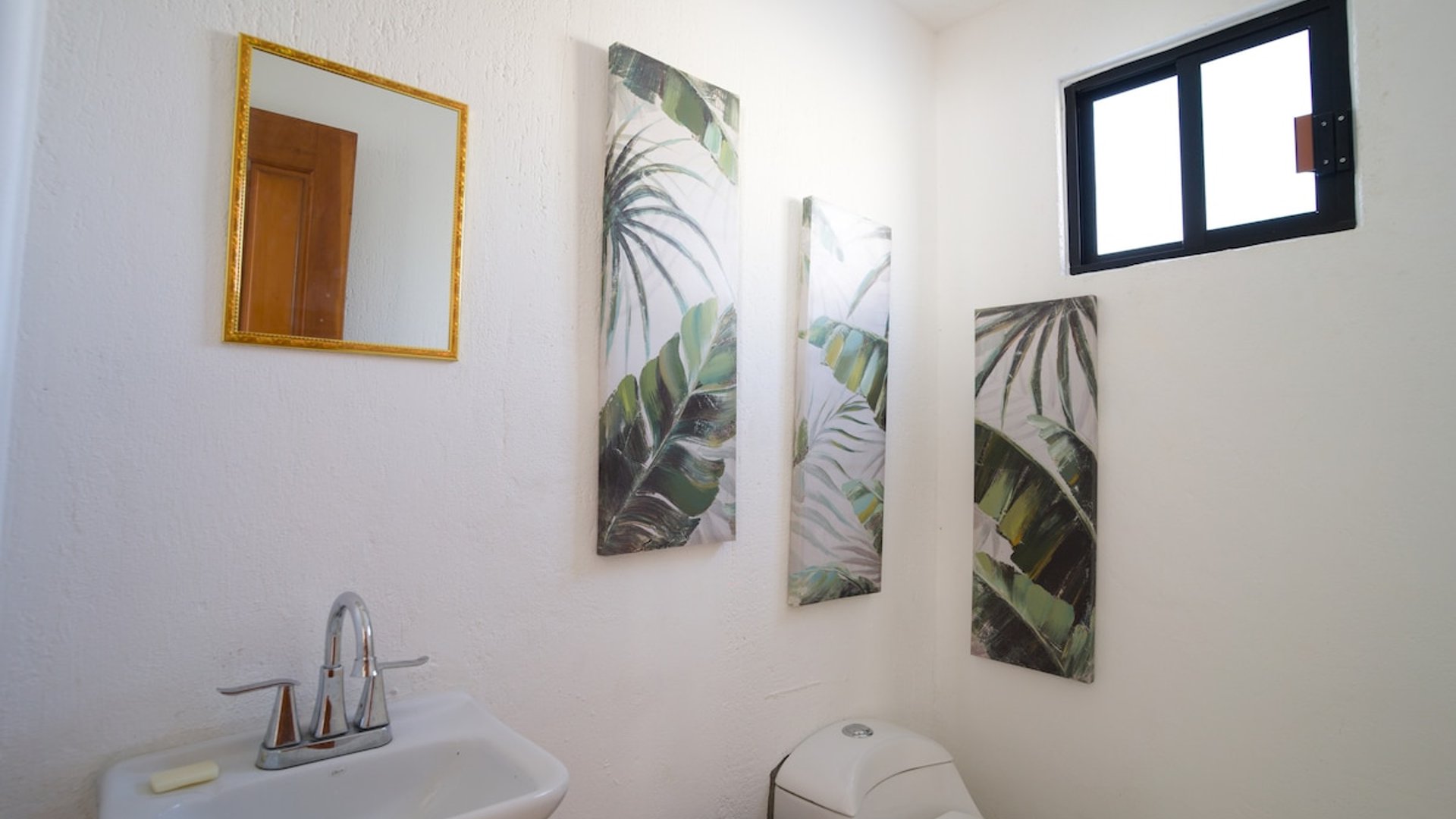 Baño blanco con lavabo, espejo dorado y tres cuadros decorativos de hojas tropicales en tonos verdes