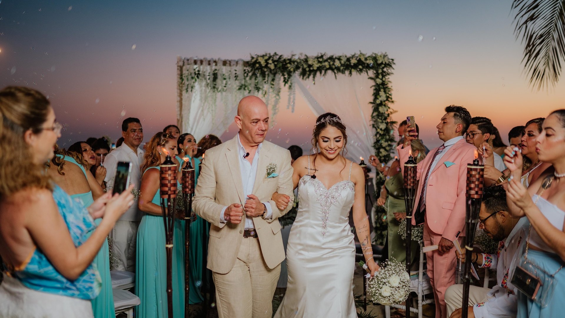 Ceremonia de boda al atardecer con arco floral decorado y vista al mar entre palmeras