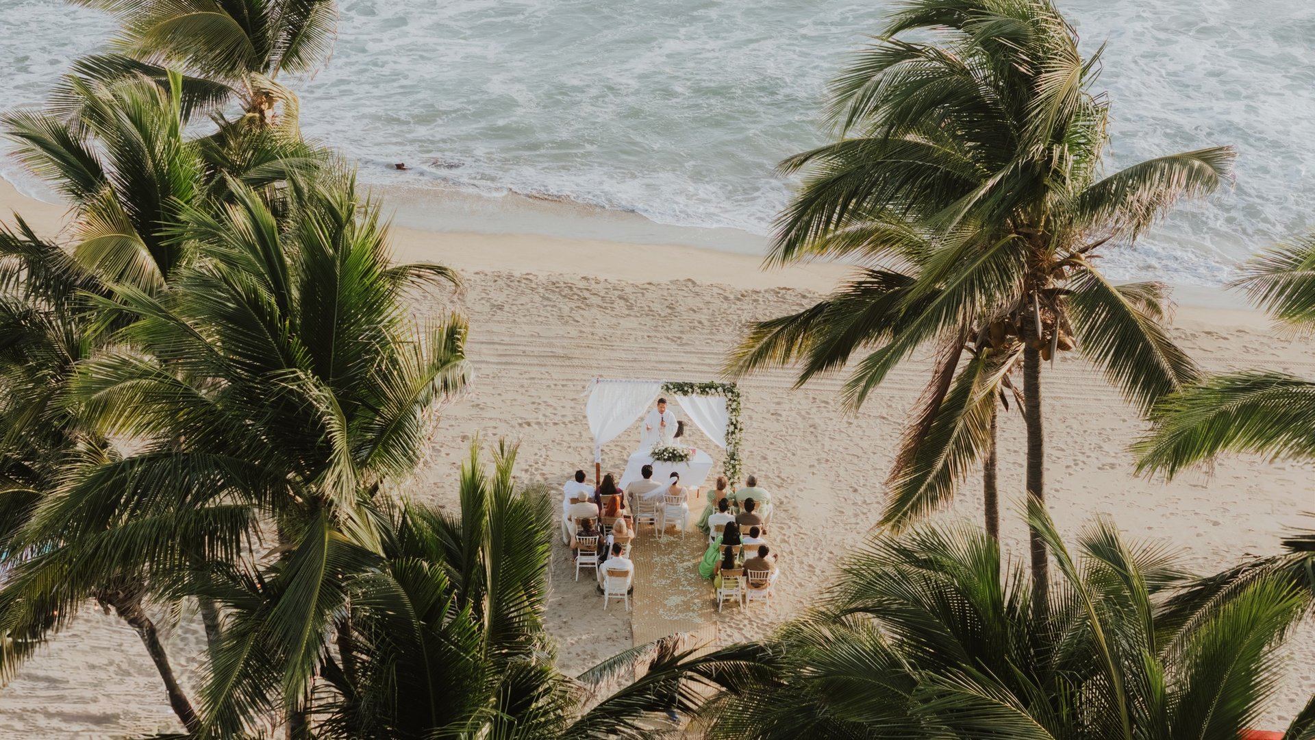 Ceremonia de boda en playa con arco blanco y sillas entre palmeras frente al Pacífico
