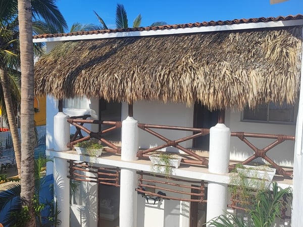 Terraza de segundo piso con techo de palapa y barandal de bambú rodeada de palmeras bajo cielo azul