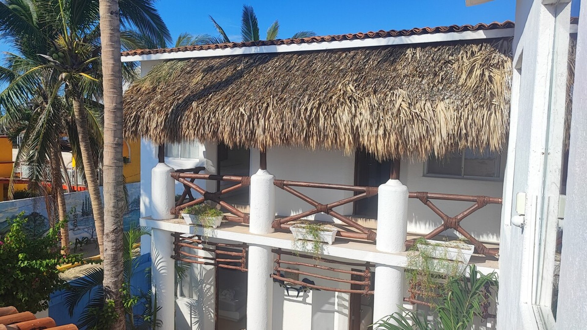 Terraza de segundo piso con techo de palapa y barandal de bambú rodeada de palmeras bajo cielo azul