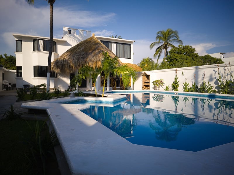 Villa blanca de dos niveles con alberca privada, palapa central y palmeras bajo cielo azul