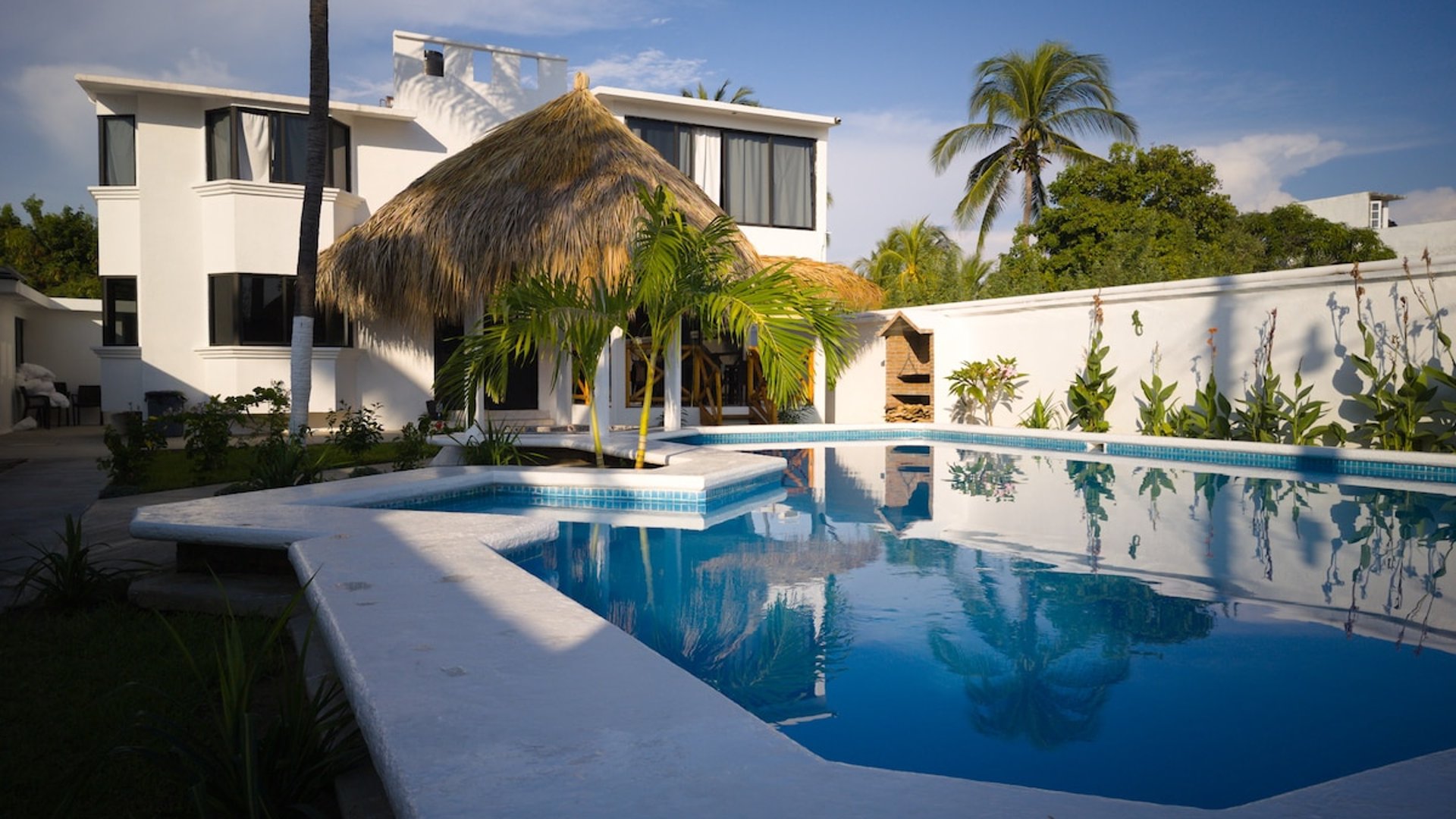 Villa blanca de dos niveles con alberca privada, palapa central y palmeras bajo cielo azul