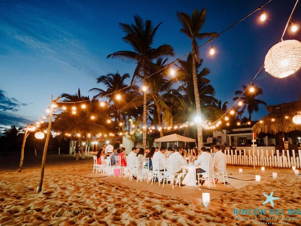 Cena de gala en la playa al anochecer con luces colgantes, mesas blancas y palmeras tropicales