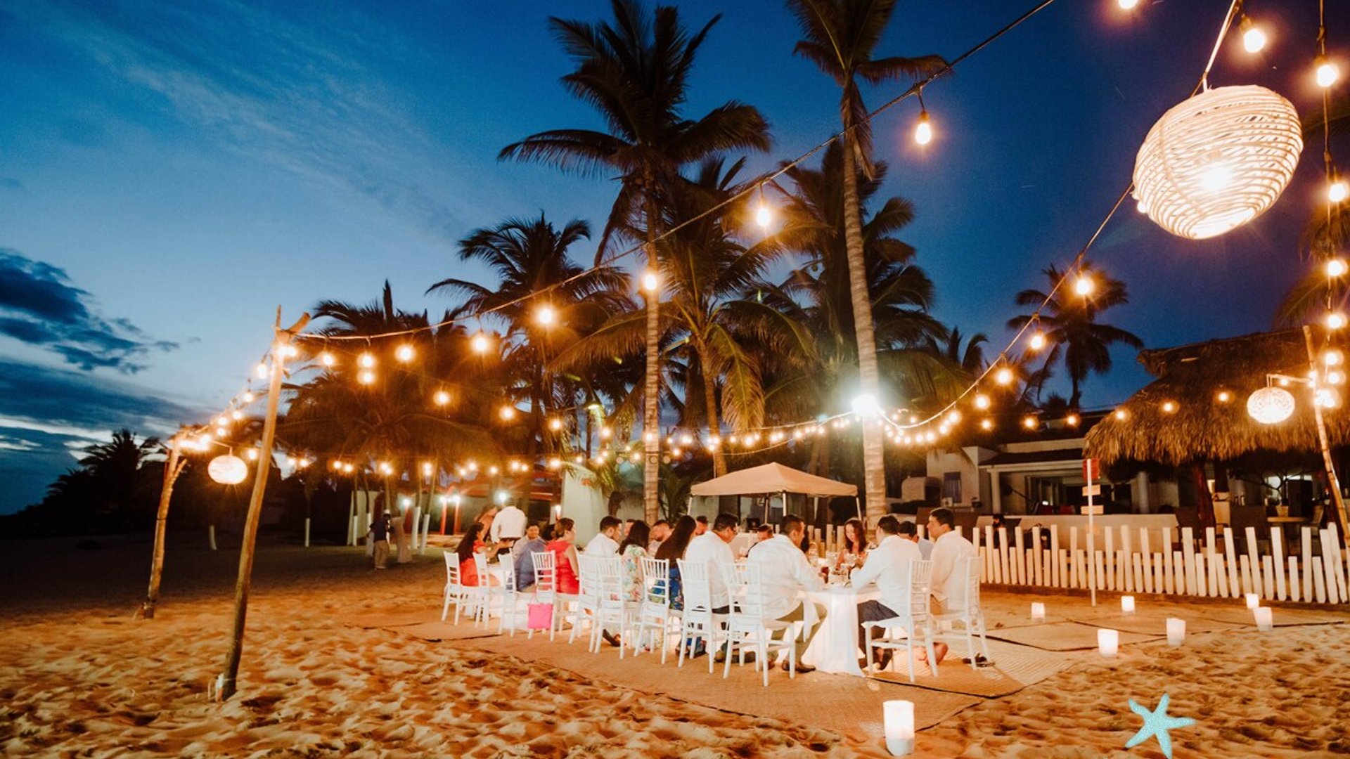 Cena de gala en la playa al anochecer con luces colgantes, mesas blancas y palmeras tropicales