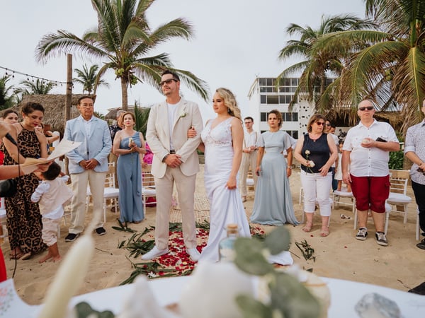 Ceremonia de boda en playa con invitados rodeando el pasillo decorado con pétalos bajo palmeras