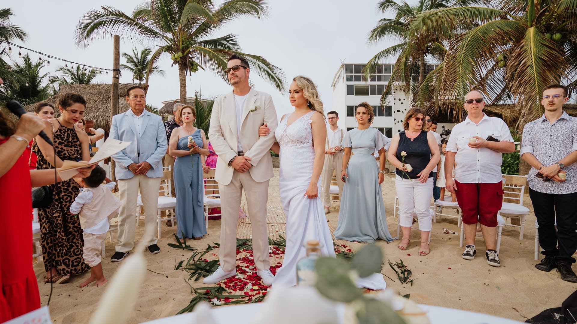 Ceremonia de boda en playa con invitados rodeando el pasillo decorado con pétalos bajo palmeras