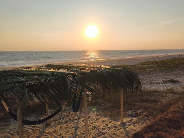 Palapa rústica con hamaca frente al mar al atardecer, sol tocando el horizonte sobre la playa desierta