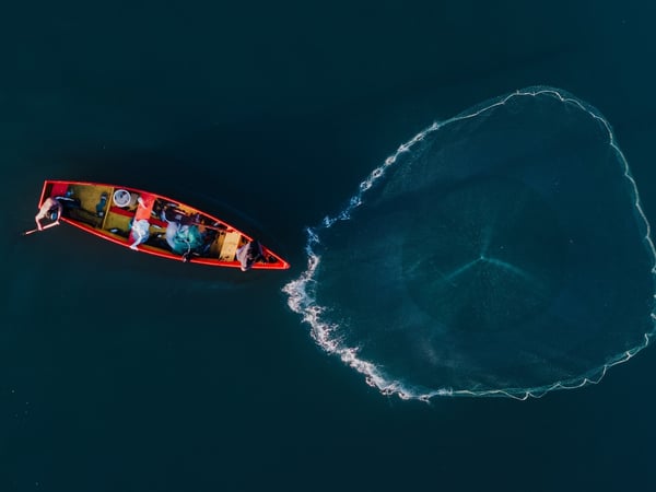 Vista aérea de pescadores en lancha roja lanzando red circular sobre aguas profundas y tranquilas